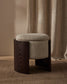 A modern storage ottoman stool with a curved dark wood frame and a round, textured light beige fabric cushion, set against a neutral background with a beige curtain and textured rug.