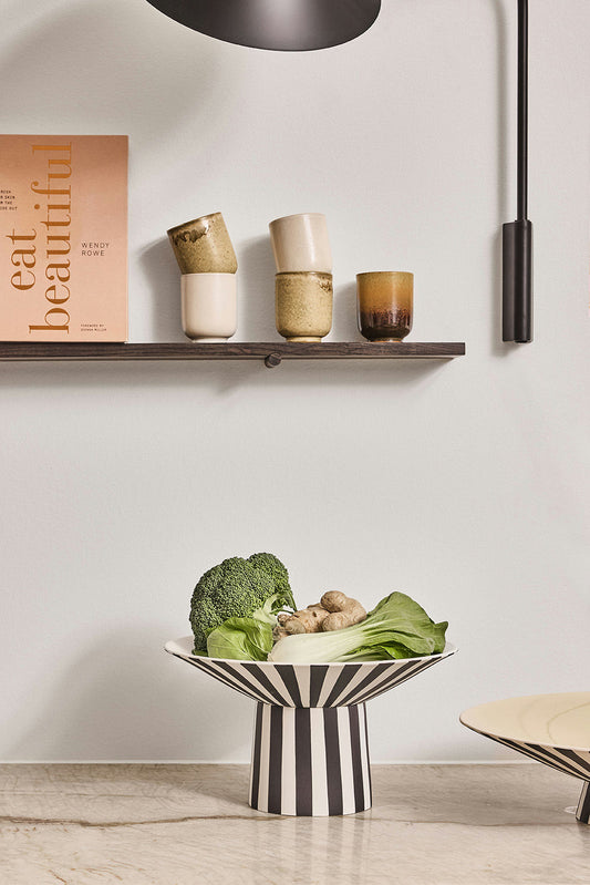 Striped ceramic bowl with vegetables on a wooden surface, with a shelf and decor items in the background.