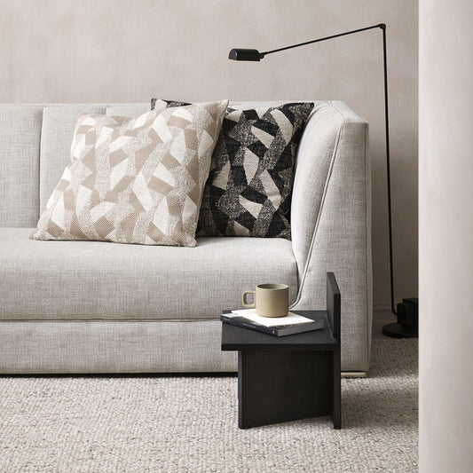 2 oscar cushions on a grey linen sofa, one is a cream and beige geometric abstract and other is black and charcoal. a small black floor lamp to the right and a little black wooden side table in front with a book and a green cup of coffee on top