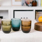 Three ribbed glass tumblers in smoky translucent shades of olive green, teal blue, and amber brown, arranged on a white textured tablecloth in a bright, modern room setting.