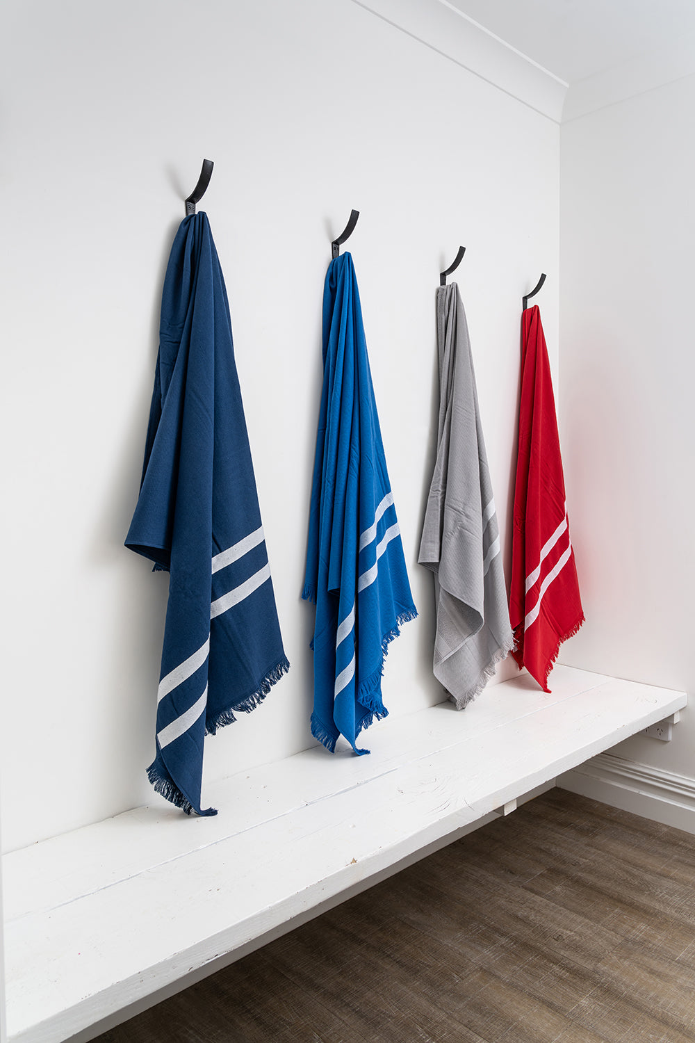 Four peshtemal towels in blue, gray, and red hanging on hooks against a white wall.