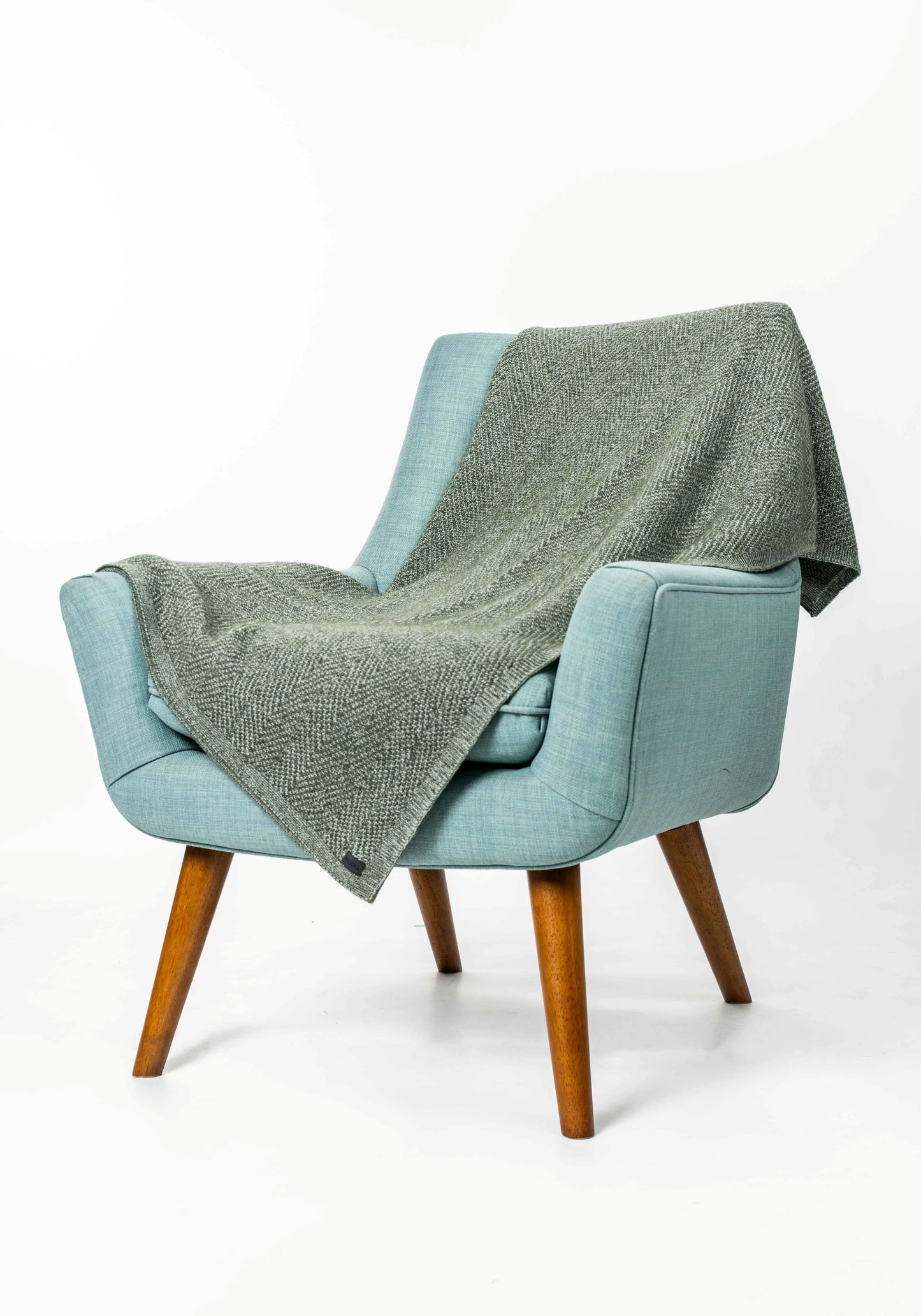 A light blue upholstered armchair with wooden legs, draped with a textured sage green throw blanket, set against a plain white studio background.