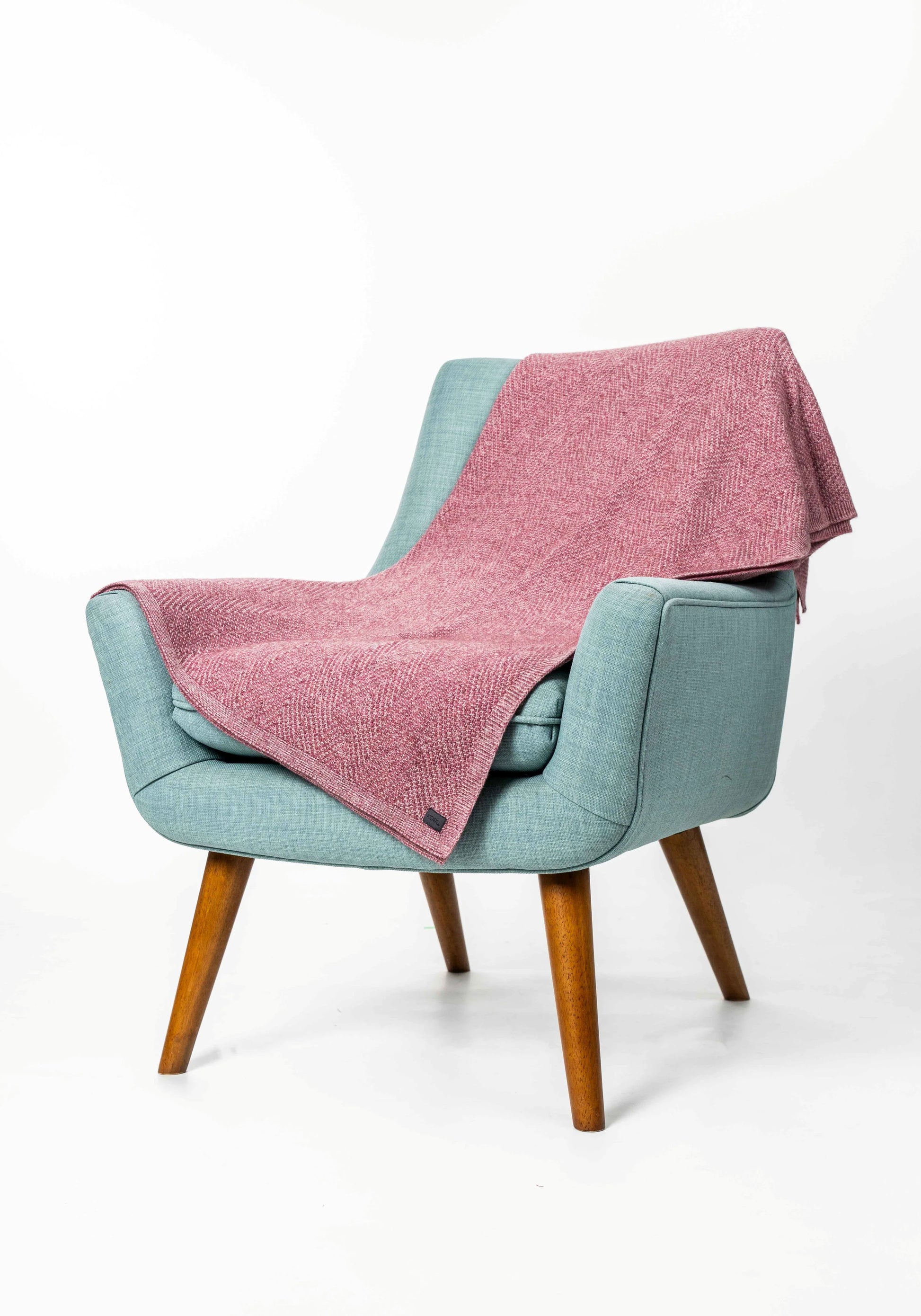 A dusty pink textured throw blanket draped over a light blue upholstered armchair with wooden legs, set against a plain white studio background.