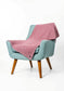 A dusty pink textured throw blanket draped over a light blue upholstered armchair with wooden legs, set against a plain white studio background.
