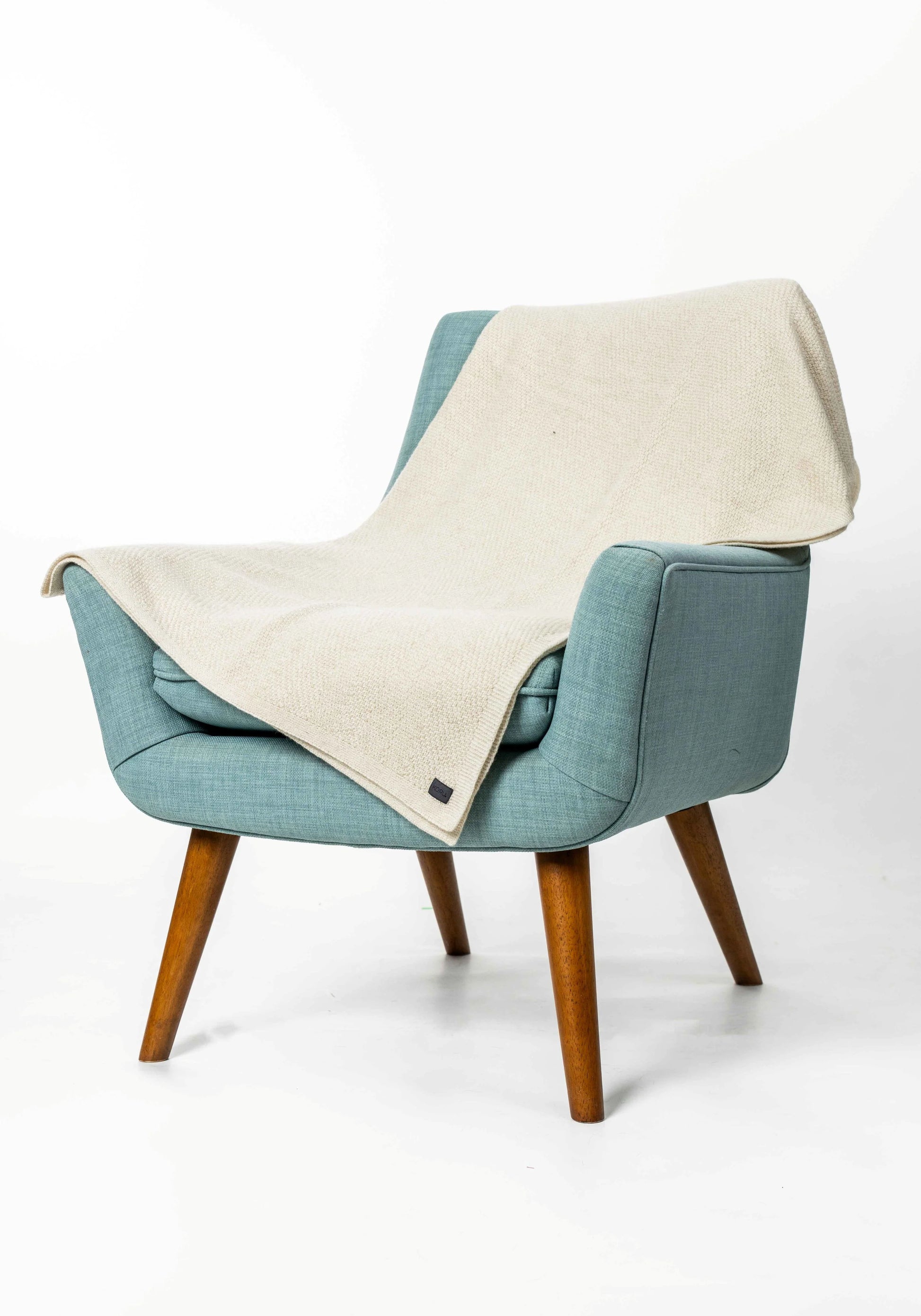 A mid-century modern armchair upholstered in textured teal fabric with tapered wooden legs, draped with a cream-colored linen throw blanket, set against a plain white studio background.
