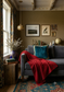 A cozy living room corner featuring a gray sofa with a bright red throw blanket draped over it, a teal velvet pillow, patterned curtains, framed wall art, and a patterned rug.