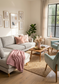A bright, modern living room featuring a light gray sectional sofa with pink throw pillows and a dusty pink blanket, a wooden coffee table, a woven rug, and a mint green accent chair near a large window.