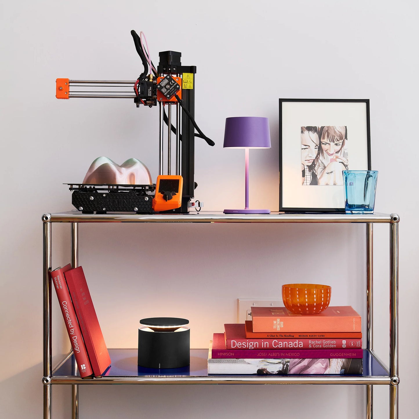 Metal shelf with a 3D printer, books, lamp, and decorative items against a light background