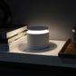 White cylindrical lamp on a dark surface with books and a glass in the background