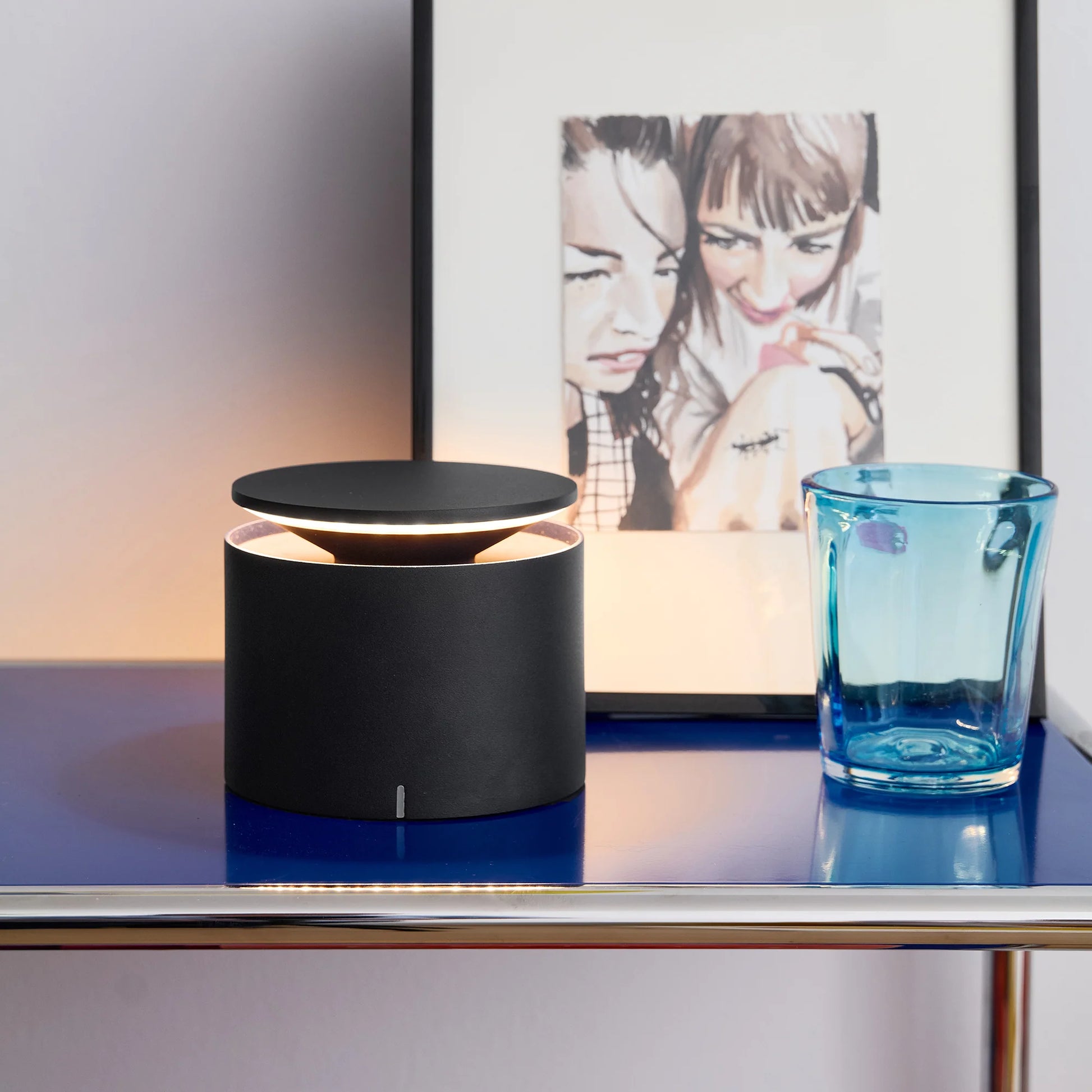 Black cylindrical lamp on a table with a blue glass and framed artwork in the background