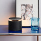 Black cylindrical lamp on a table with a blue glass and framed artwork in the background