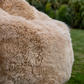 A close-up of a fluffy, shaggy beige outdoor bean bag chair on a green lawn with blurred foliage in the background.