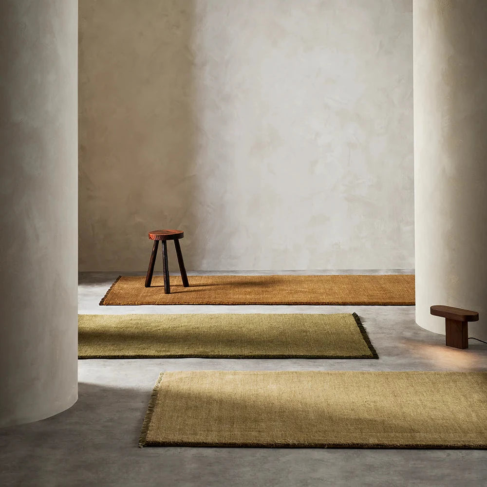 3 runner rugs in brown, green and beige on a stone floor with stone walls and two stone pillars with a wooden stool sitting on the brown rug
