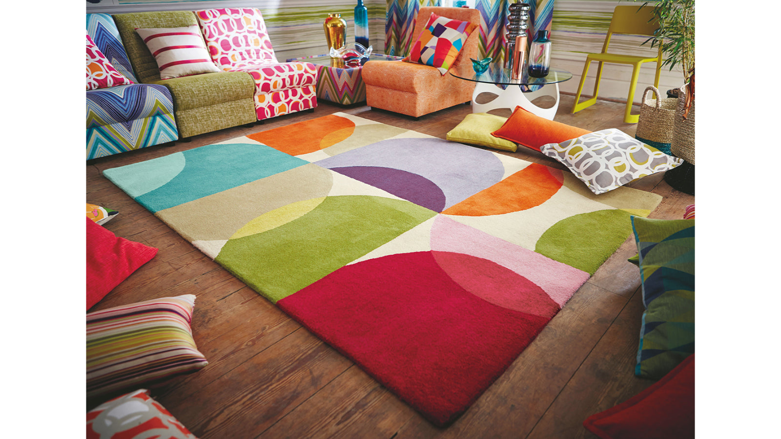 A colorful area rug with large abstract semicircle shapes in red, teal, green, orange, purple, and cream, styled in a modern living room setting on a dark wood floor.