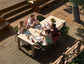 Group of people sitting around a table on a wooden deck, enjoying a meal together.