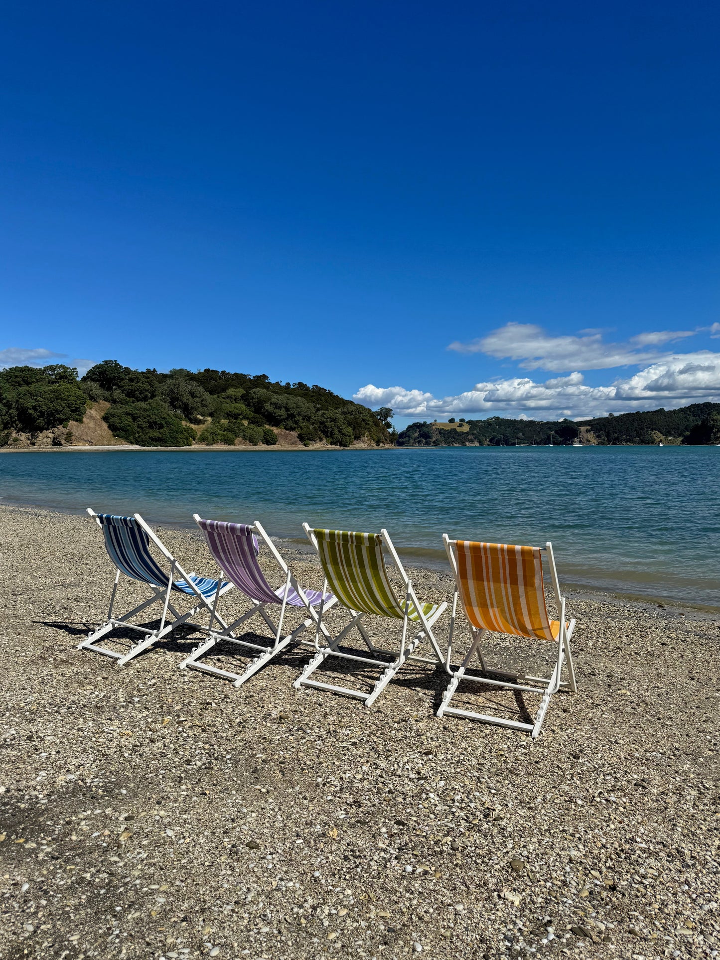 Colorful beach chairs on a pebbly beach with a body of water and trees in the background.