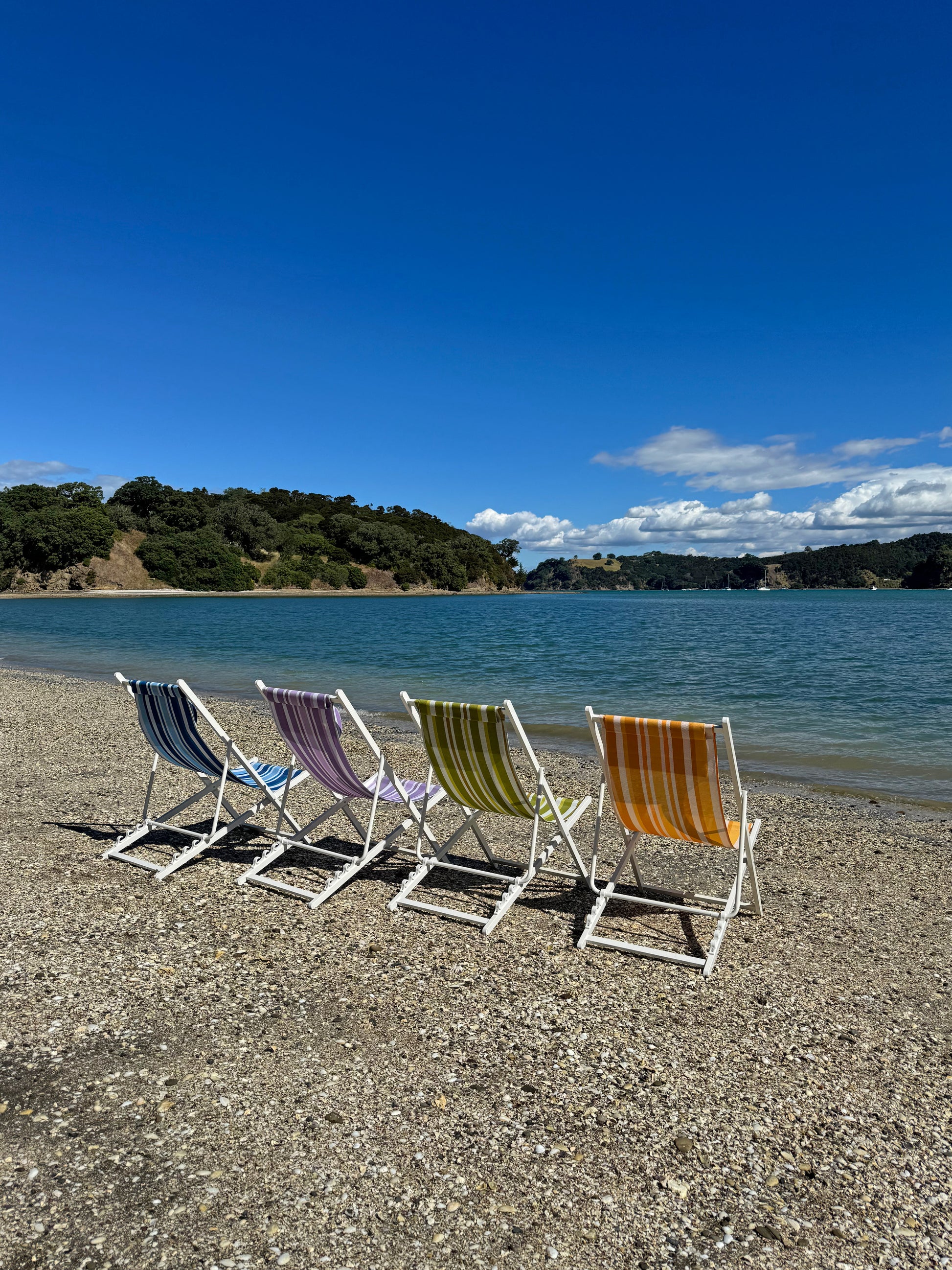 Colorful beach chairs on a pebbly beach with a body of water and trees in the background.
