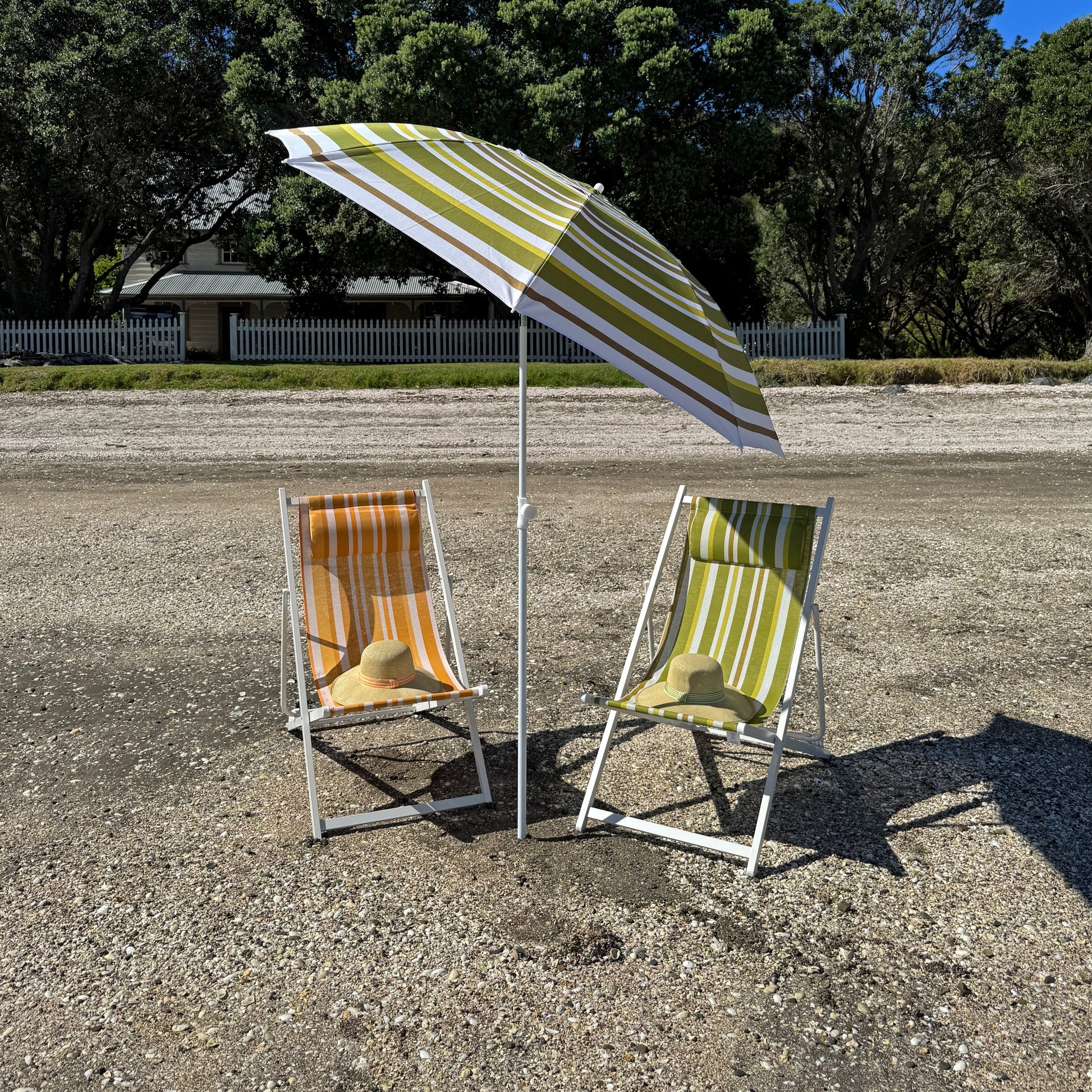 Two beach chairs under a striped umbrella on a gravel surface with trees in the background.
