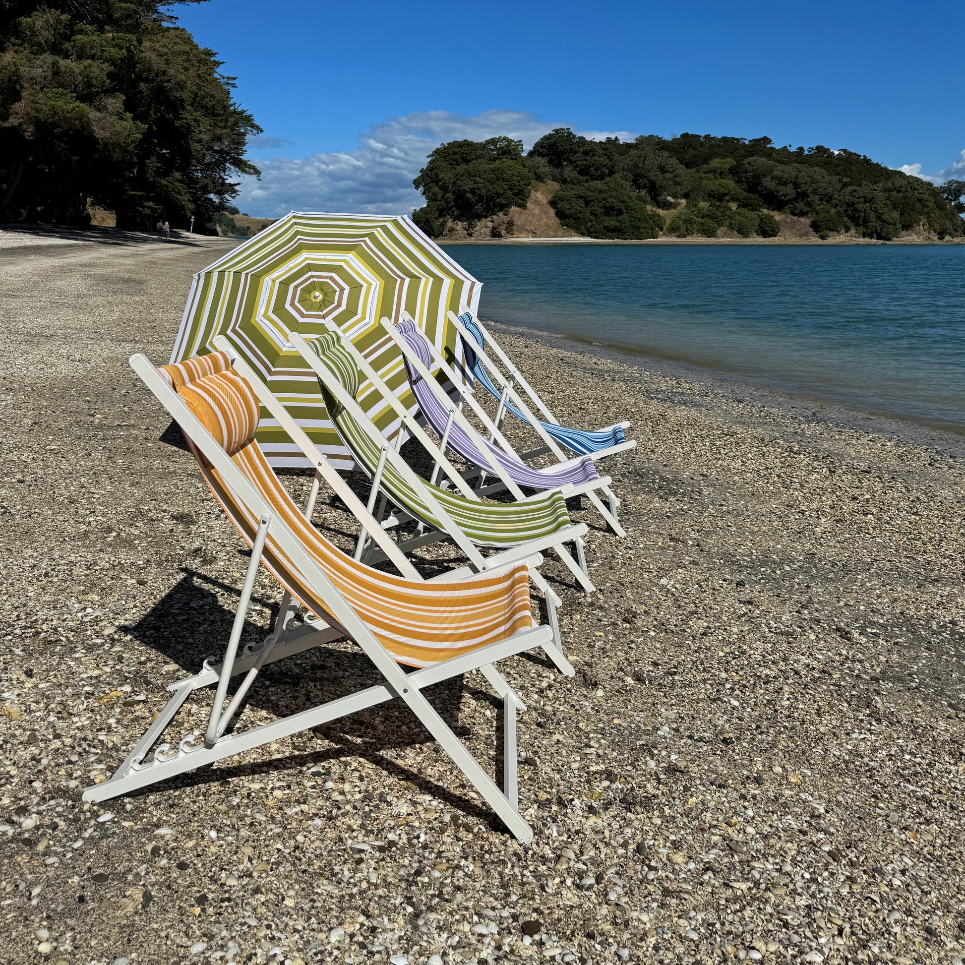 Beach chairs and an umbrella on a pebbly beach with trees and water in the background.