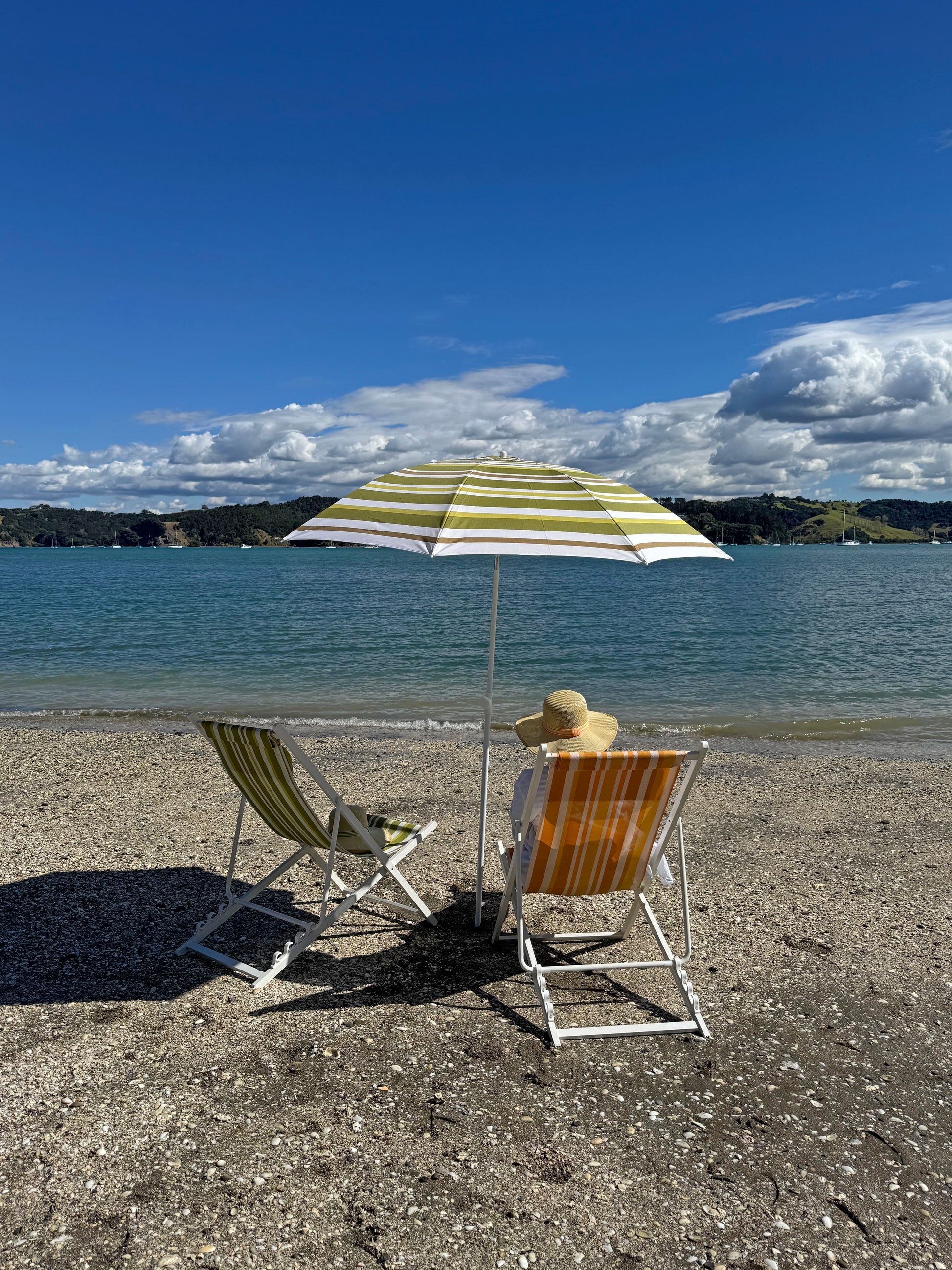 Two beach chairs with a striped umbrella on a sandy beach with a scenic view of water and sky.