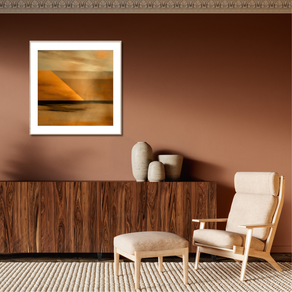 A framed abstract landscape art print with warm orange, yellow, and brown tones hangs on a terracotta-colored wall above a dark wood sideboard in a styled living room setting with a beige armchair and ottoman.