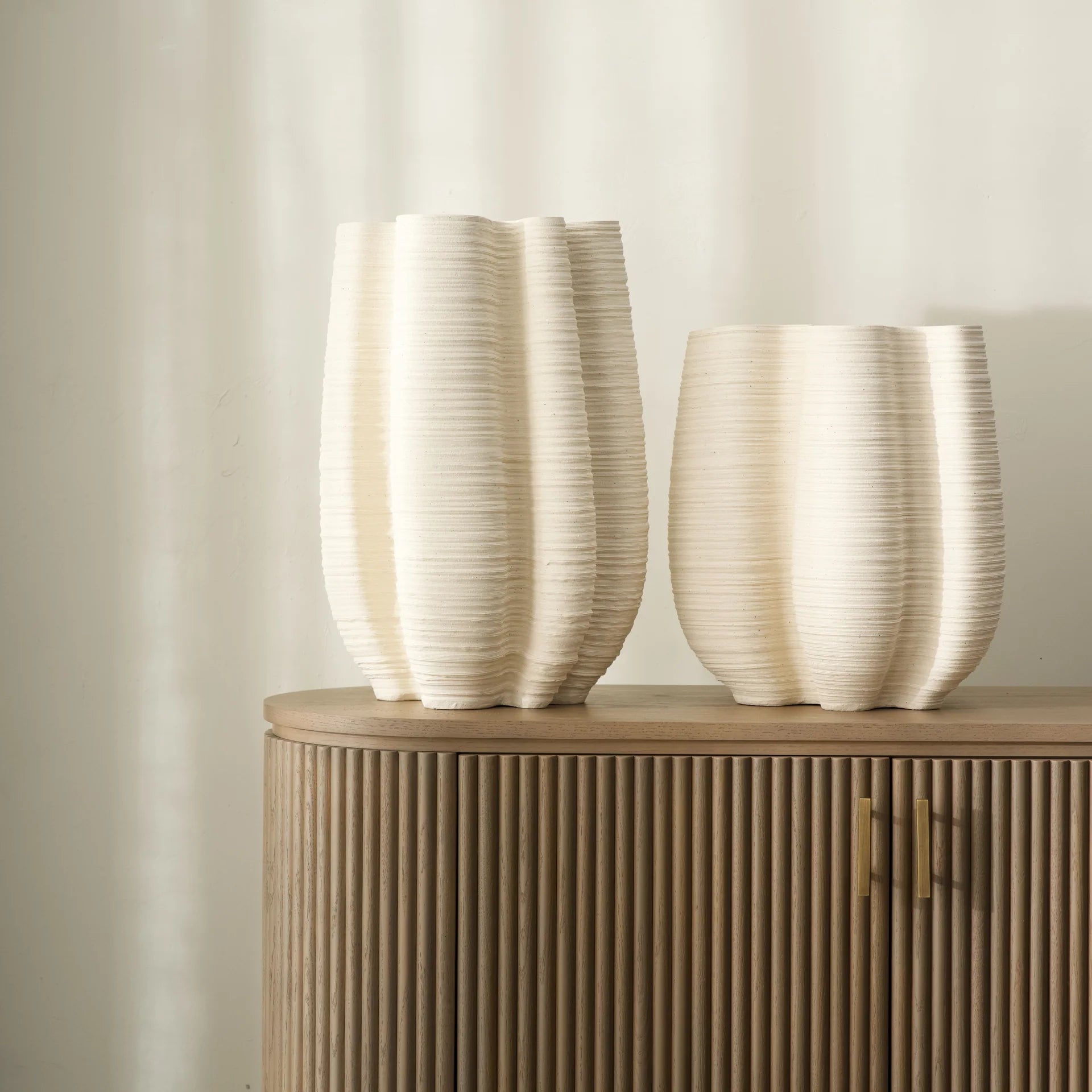 Two white textured vases on a wooden cabinet against a beige wall