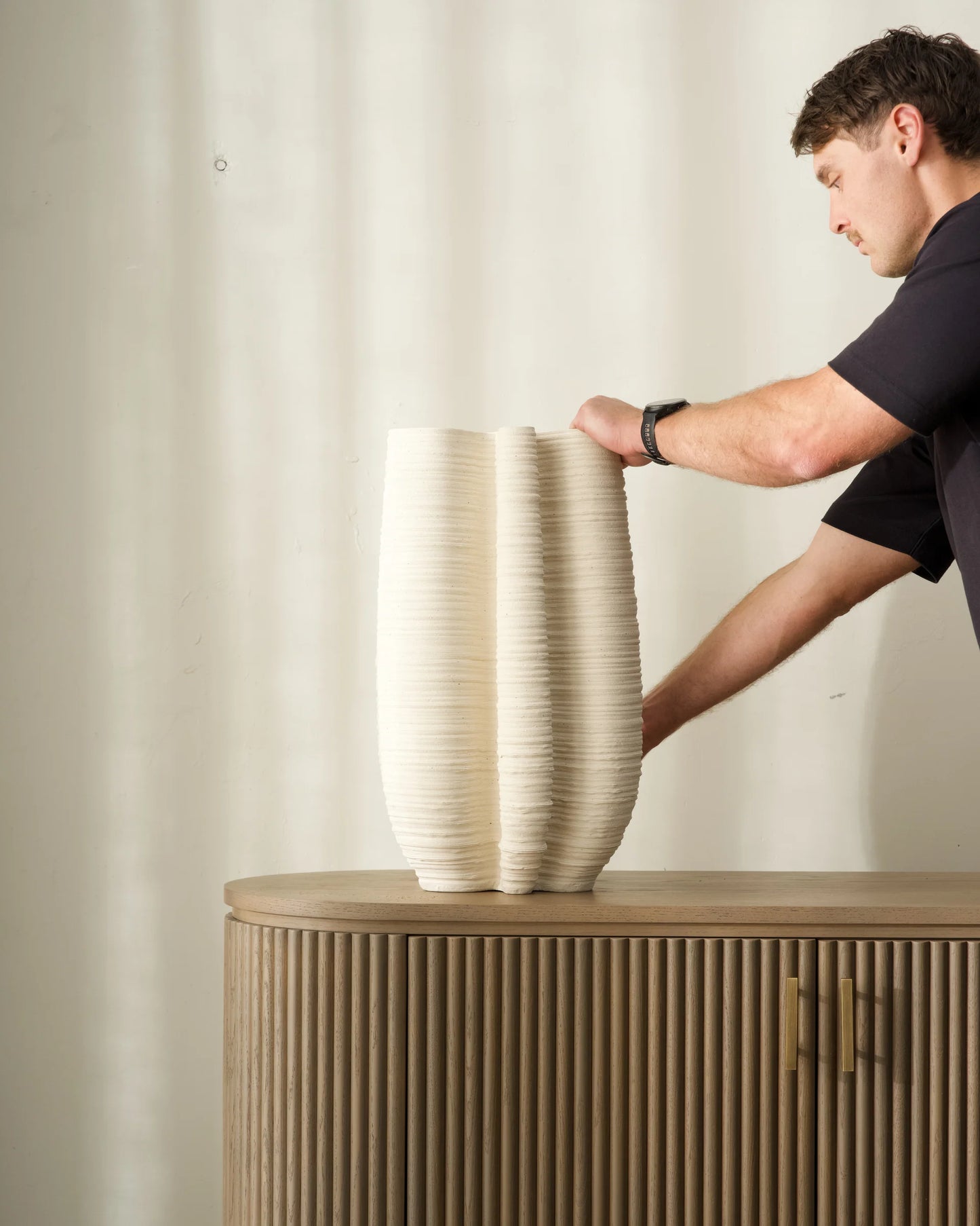 Man placing a large decorative vase on a wooden cabinet against a plain wall.