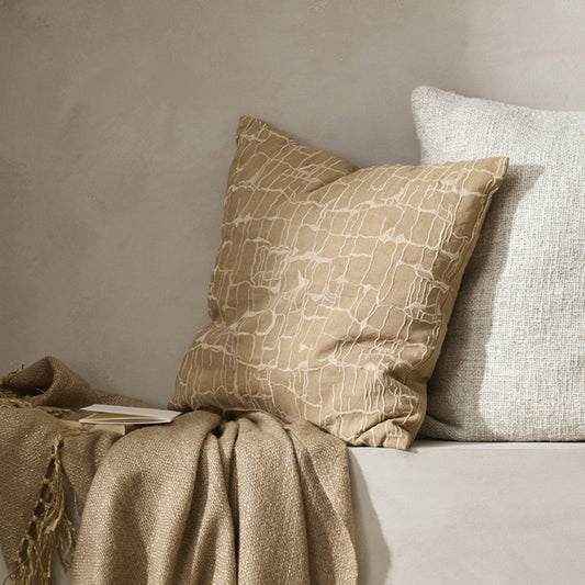 Triton cushion in a peachy colour with white embroidered lines sitting against a cream textured cushion on a white plastered shelf with a beige folded thrown in front