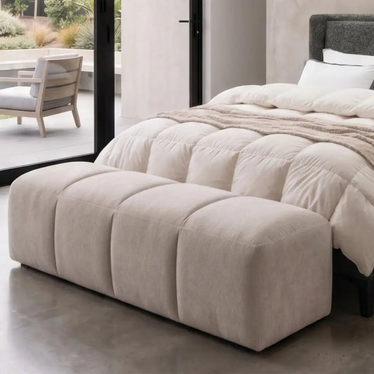 Cream end of bed ottoman in 4 padded sections by a cream quilted bed and concrete floor with black framed doors viewing onto a patio with furniture
