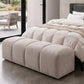 Cream end of bed ottoman in 4 padded sections by a cream quilted bed and concrete floor with black framed doors viewing onto a patio with furniture
