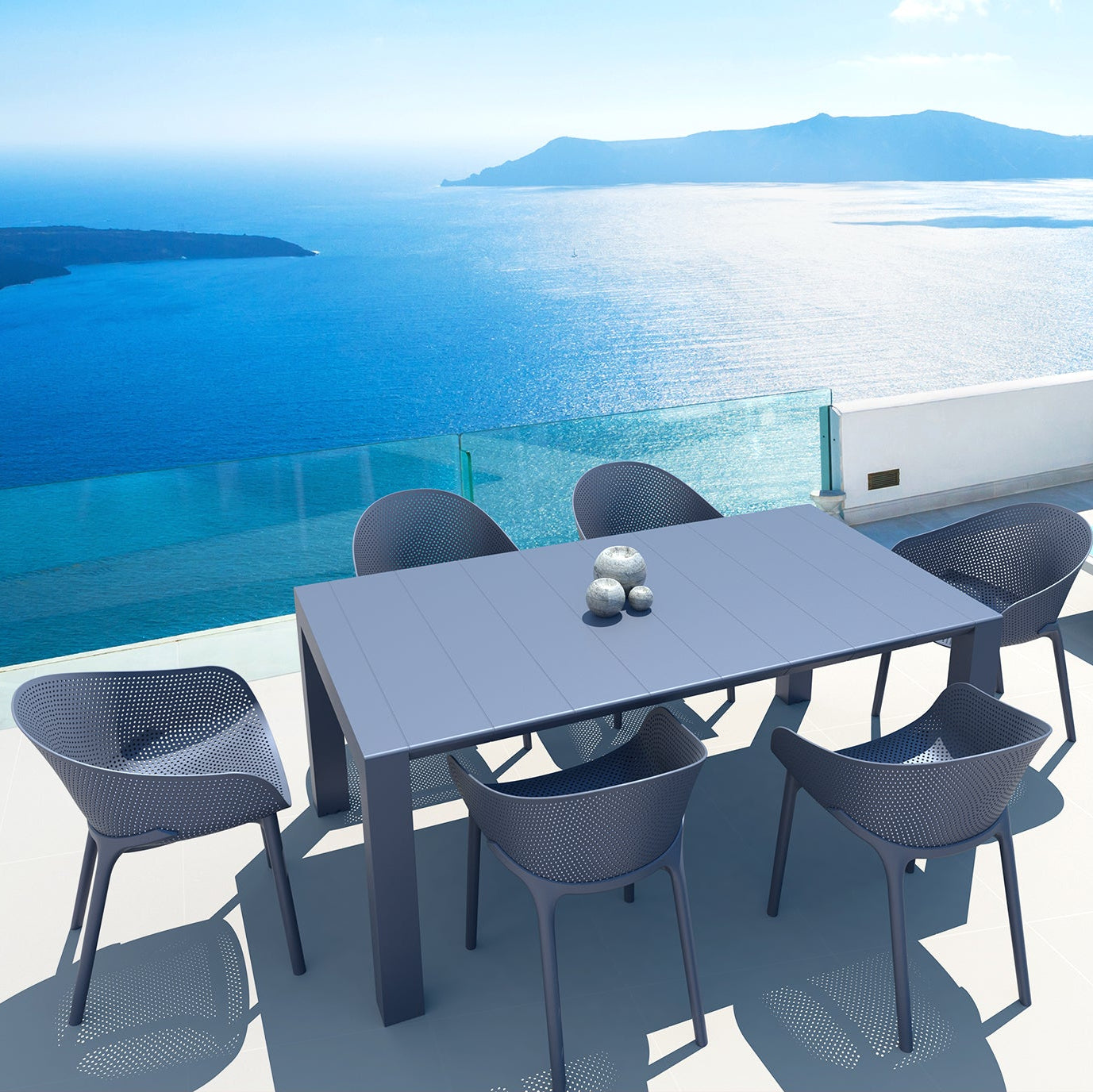 Outdoor dining table with chairs on a balcony overlooking a blue sea and mountains.