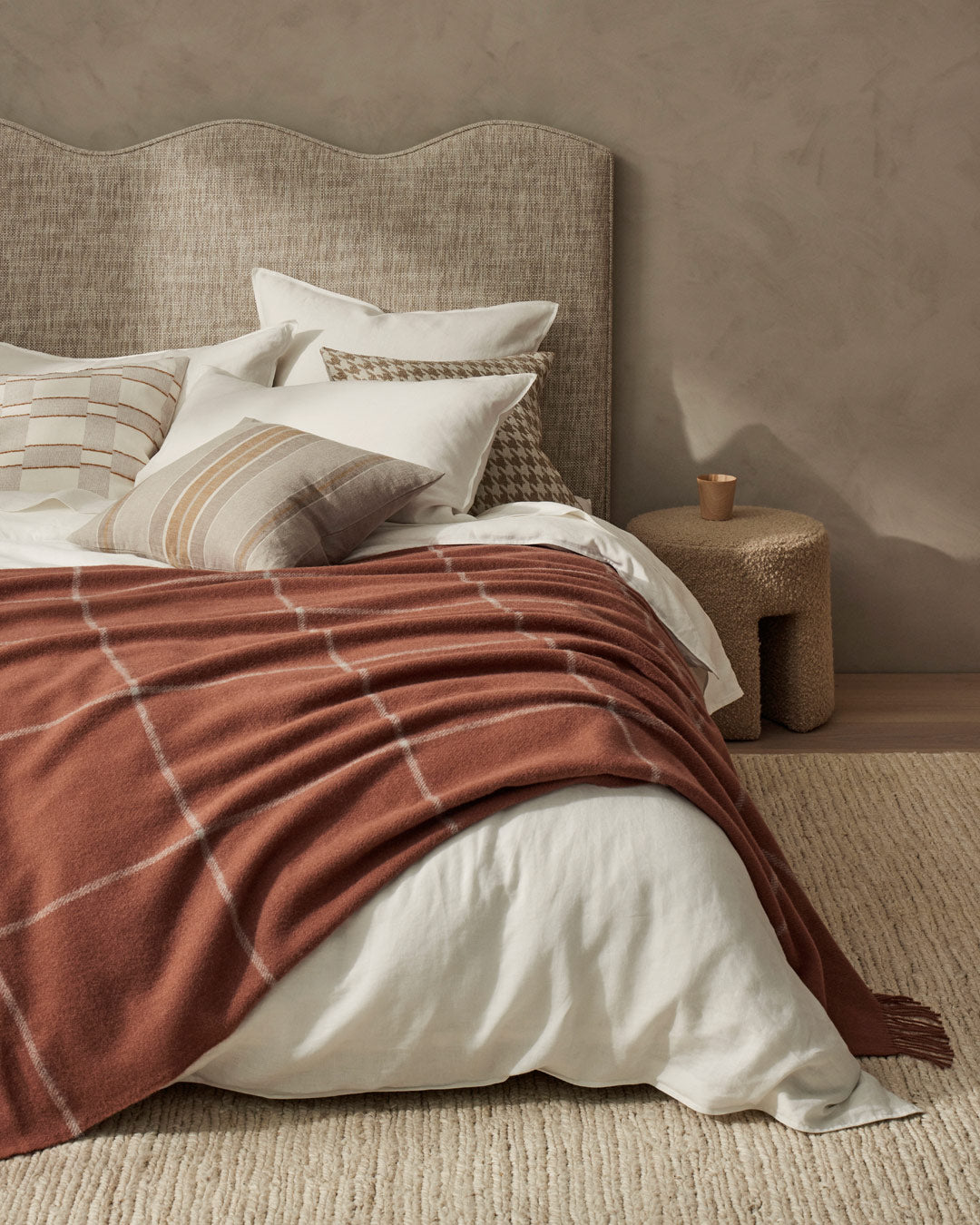 warm toned ranfurly throw with large scale check  draped over a bed with white linen and beige walls and head board. Sitting on a pale cream wool carpet with neural colours on the beds cushion. Along sie is a small boucle fabric side table