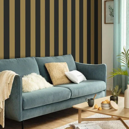 A modern living room scene featuring a prominent wall with vertical stripes in black and a warm mustard-gold color. In front of the wall is a teal-blue velvet sofa styled with white and beige throw pillows, a cream throw blanket, and a light wood coffee table in the foreground.