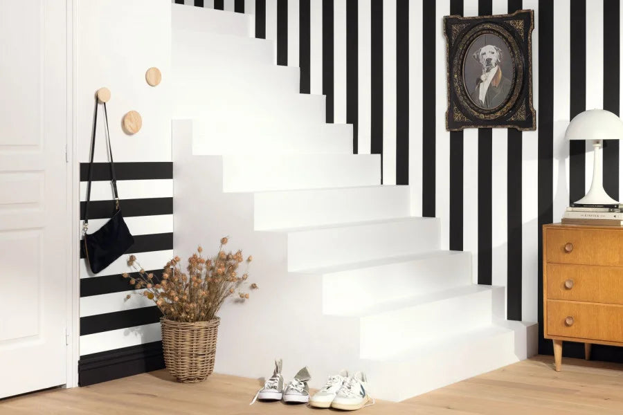 A modern staircase with white steps and bold black-and-white vertical stripes on the walls, styled with a wicker basket of dried flowers, a black shoulder bag on a wall hook, white sneakers at the base, a wooden cabinet, and a framed portrait.