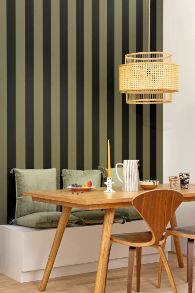 A modern dining area featuring a wooden table and chair in front of a wall with vertical olive green and black stripes. A white bench with green cushions sits underneath, and a woven rattan pendant light hangs from the ceiling.