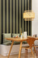 A modern dining area featuring a wooden table and chair in front of a wall with vertical olive green and black stripes. A white bench with green cushions sits underneath, and a woven rattan pendant light hangs from the ceiling.