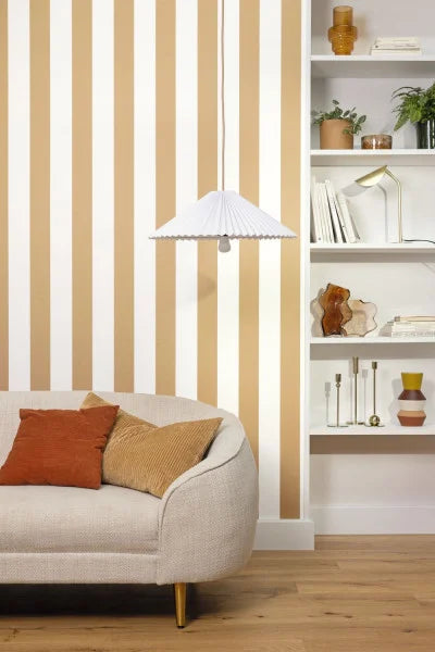 A styled interior scene showing a cream upholstered sofa with rust and tan pillows, a white hanging pendant lamp, beige-and-white vertically striped wallpaper, light wood flooring, and a white built-in bookshelf with decor.
