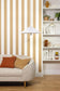 A styled interior scene showing a cream upholstered sofa with rust and tan pillows, a white hanging pendant lamp, beige-and-white vertically striped wallpaper, light wood flooring, and a white built-in bookshelf with decor.