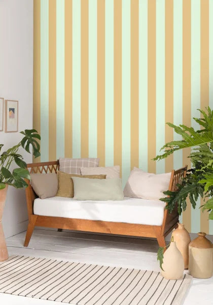 A styled interior scene featuring a wooden daybed with a white cushion and neutral pillows, set against a wall with vertical stripes in pale green and warm beige. The room includes potted green plants, beige ceramic vases, and a striped rug.
