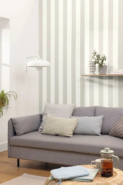 A modern gray fabric sofa in a bright living room, styled with neutral throw pillows, a striped pillow, a wall lamp, a floating shelf with a small plant, and a coffee table in the foreground.