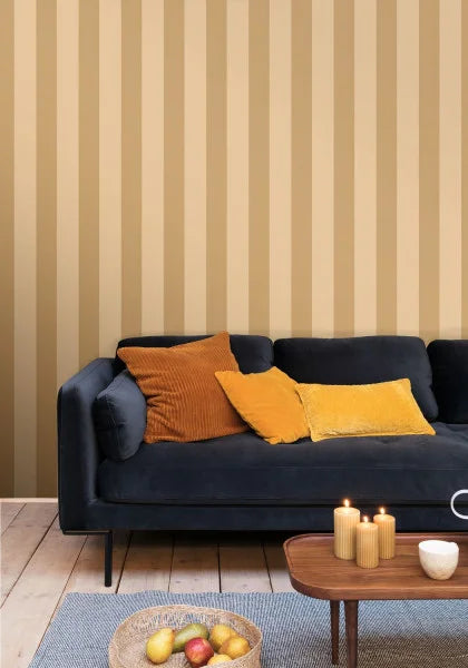 A styled living room scene featuring a dark navy blue velvet sofa with mustard yellow and orange-brown accent pillows, set against a beige and brown vertically striped wallpaper. A wooden coffee table with lit candles and a bowl of fruit sits on a blue rug in the foreground.
