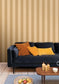 A styled living room scene featuring a dark navy blue velvet sofa with mustard yellow and orange-brown accent pillows, set against a beige and brown vertically striped wallpaper. A wooden coffee table with lit candles and a bowl of fruit sits on a blue rug in the foreground.