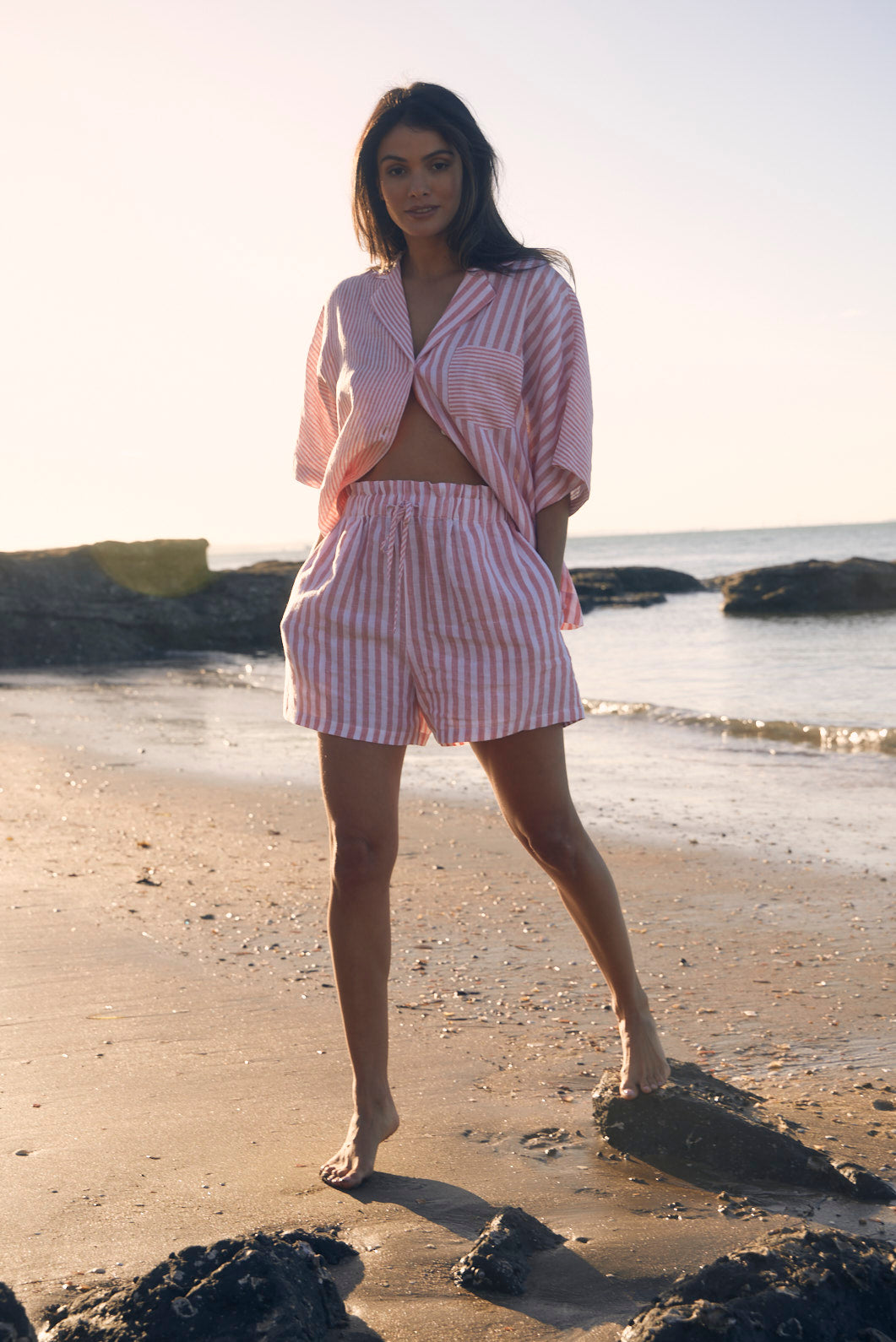 Woman in a pink striped outfit standing on a beach with water and rocks in the background.