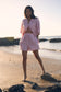 Woman in a pink striped outfit standing on a beach with water and rocks in the background.