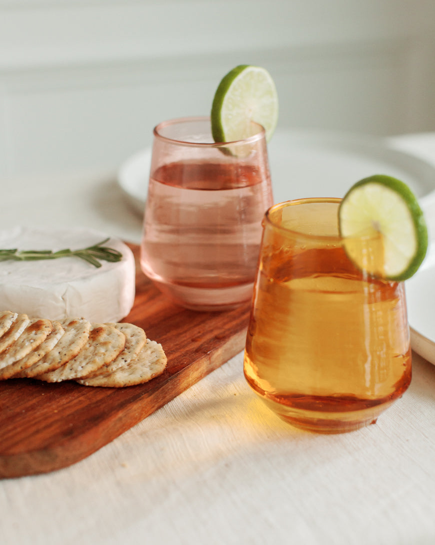 Two glasses of pink and orange drinks with lime slices on a wooden board with crackers and cheese.