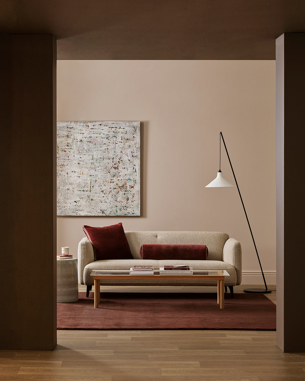 A modern living room scene featuring a beige sofa with two dark red throw pillows, a low wooden coffee table, a textured red rug, and a black floor lamp against a warm beige wall.