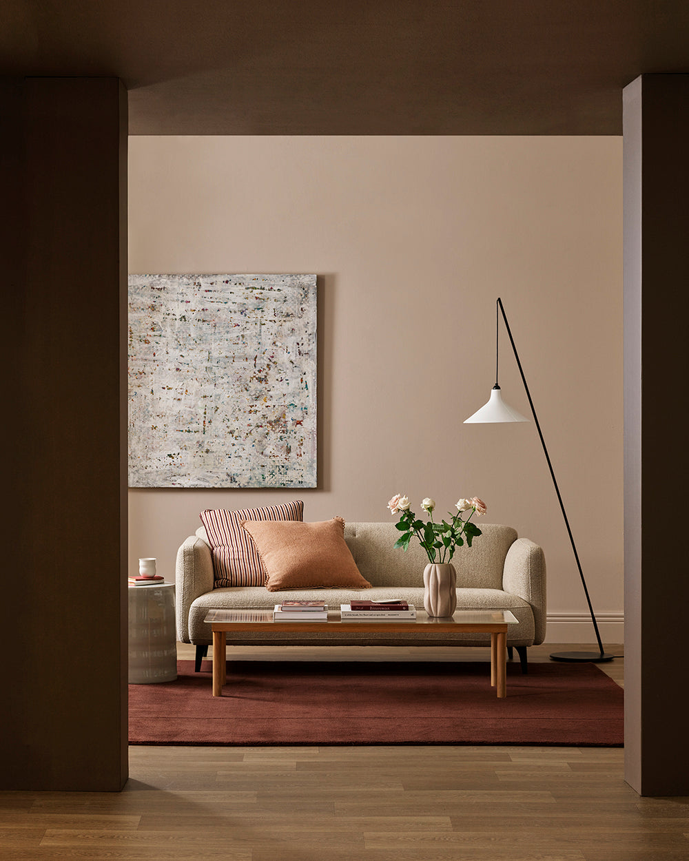 A styled living room scene viewed through a dark doorway, featuring a beige sofa with pink and striped pillows, a wooden coffee table, a vase of flowers, a floor lamp, and a large abstract painting on a warm beige wall.