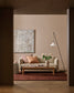 A styled living room scene viewed through a dark doorway, featuring a beige sofa with pink and striped pillows, a wooden coffee table, a vase of flowers, a floor lamp, and a large abstract painting on a warm beige wall.