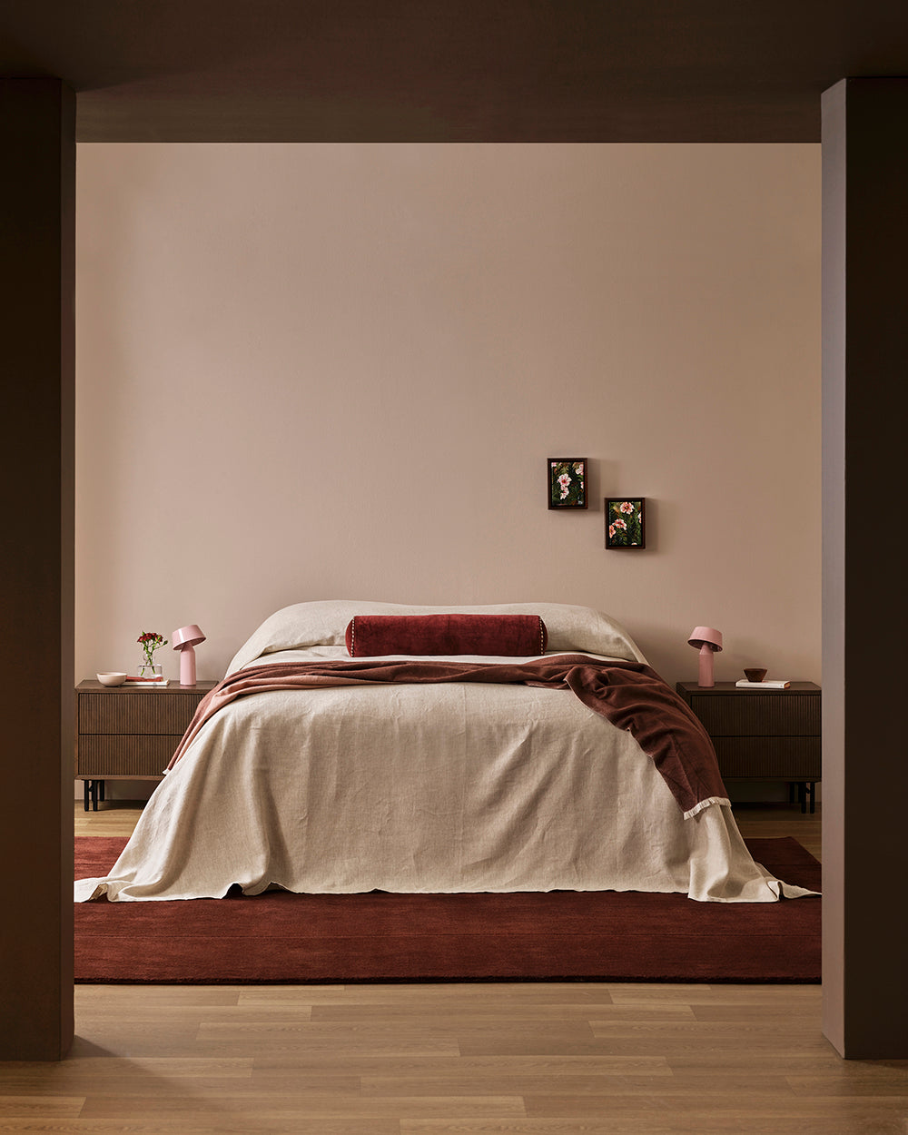 A modern bedroom scene featuring a bed dressed in cream linen bedding with a dark red cylindrical pillow and a draped dark red throw blanket. The bed is set against a warm beige wall with two small framed pictures, flanked by dark wood nightstands with pink lamps, all framed by dark brown pillars in the foreground.
