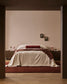 A modern bedroom scene featuring a bed dressed in cream linen bedding with a dark red cylindrical pillow and a draped dark red throw blanket. The bed is set against a warm beige wall with two small framed pictures, flanked by dark wood nightstands with pink lamps, all framed by dark brown pillars in the foreground.